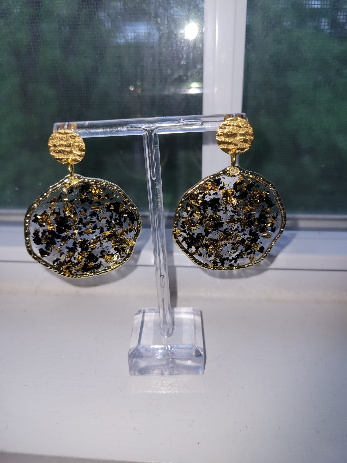 A pair of gold-toned hoop earrings with gold and black foil insert, displayed on a clear stand in front of a window.