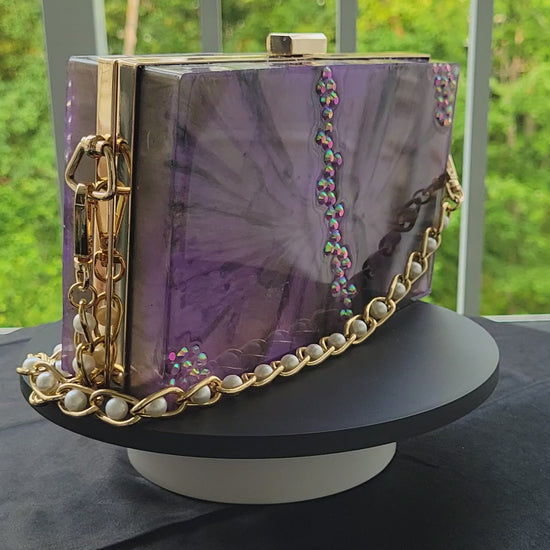 Purple, black, and white resin purse with purple jewels. Gold hardware and gold strap with pearl embellishments. Clutch is being displayed on top of a rotating display stand.