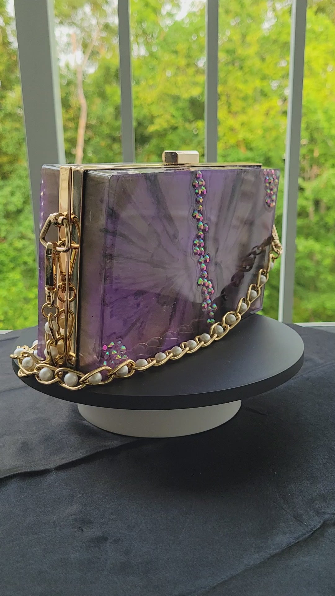 Purple, black, and white resin purse with purple jewels. Gold hardware and gold strap with pearl embellishments. Clutch is being displayed on top of a rotating display stand.