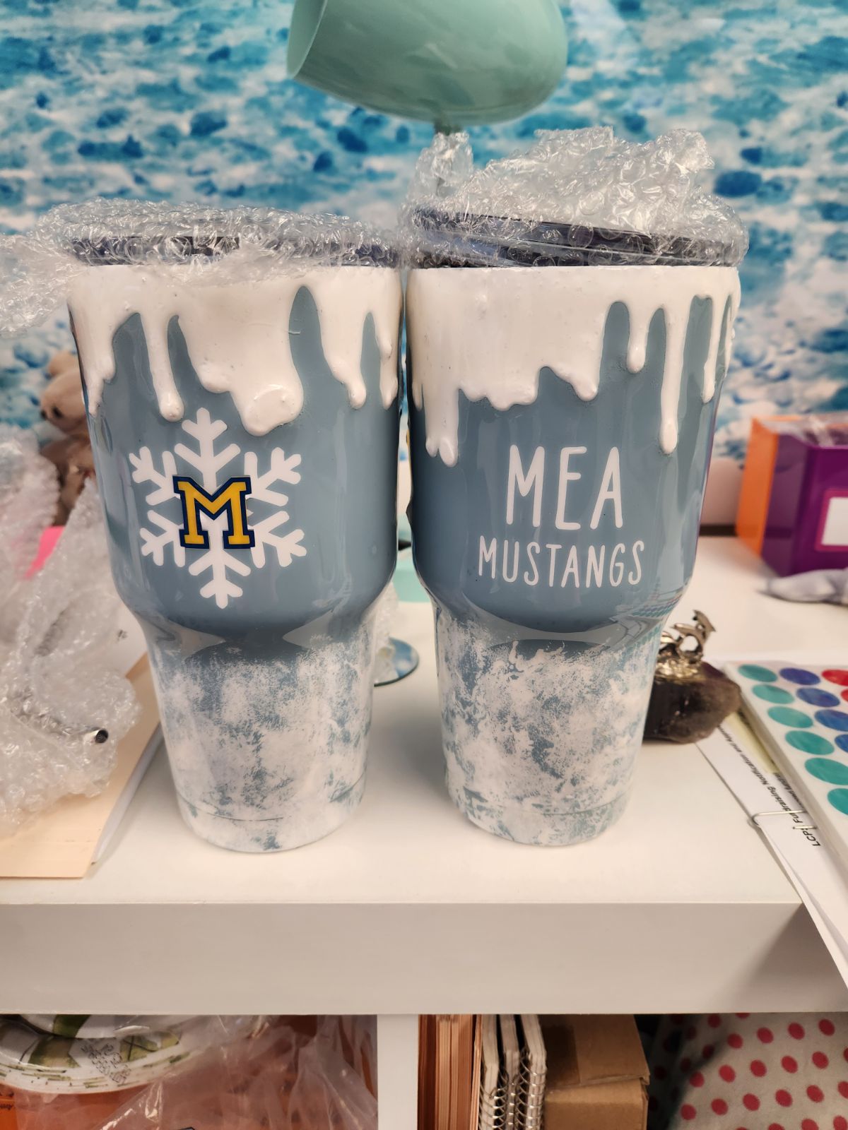 Two personalized blue tumblers with white text and blue snowflake designs, one with 'MEA MUSTANGS' and the other with a name. The tumblers are on a table with a blurred background.